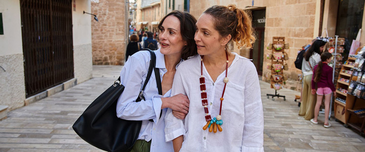 Zwei Frauen erkunden historische Bauwerke in einer mediterranen Gasse. Warme Farben und alte Architektur prägen die Atmosphäre vergangener Zeiten.